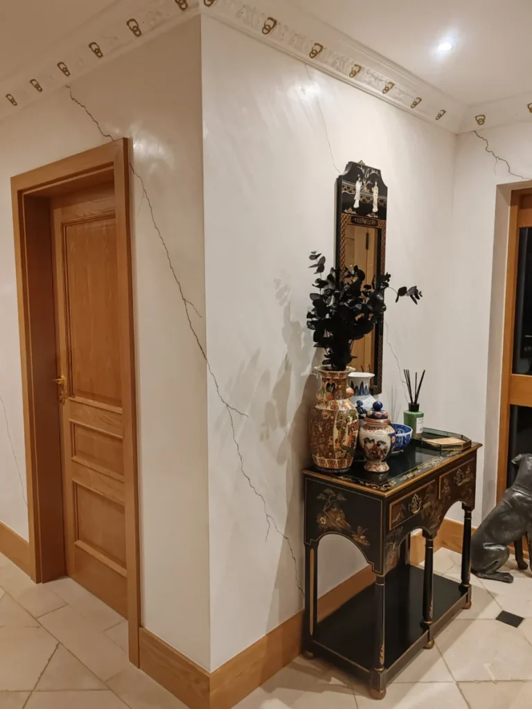 Elegant interior hallway with beige marble‑effect walls featuring decorative cracks, two wooden doors with gold handles, and a black lacquered console table with Asian‑inspired motifs. The table holds ornate ceramic vases, a blue and white bowl, and a reed diffuser. A tall mirror with a black and gold frame hangs above, and white ceiling molding with gold accents completes the refined look.